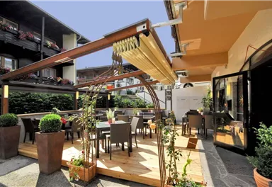 A cozy outdoor area of a restaurant with a wooden terrace and tables. Surrounded by plants and a clear sky.