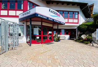 A building with the inscription "Kaiserbad" and a large entrance door.
In front of the entrance are paved paths and some plantings.