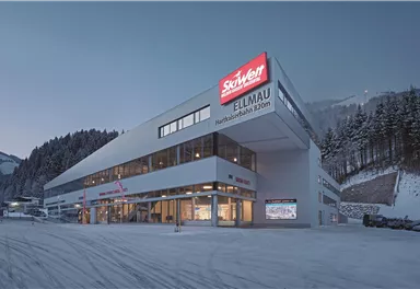 A modern building with large windows and a red sign that shows "Skisport Ellmau." It is located in a snowy environment, surrounded by trees and mountains.