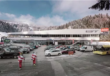 A modern building in the mountains, surrounded by snow-covered trees. In the foreground, many cars are parked and some skiers are heading towards the entrance.