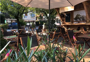 Ein gemütlicher Außenbereich mit Holztischen und Stühlen unter einem Sonnenschirm. Umgeben von grünen Pflanzen und Bäumen, bietet es einen einladenden Platz zum Entspannen.