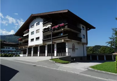 A modern building in alpine style with balconies and flowers. The surroundings are green and sunny, overlooking the mountains.