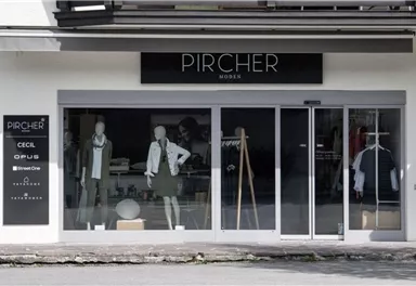 Ein moderner Bekleidungsladen mit Schaufensterpuppen, die stilvolle Kleidung präsentieren. Der Laden trägt den Namen "PIRCHER" und hat eine elegante Fassadengestaltung.