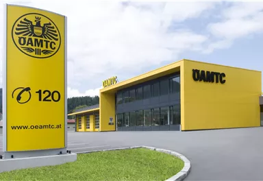 A modern building of the ÖAMTC with a striking yellow facade. In the foreground is a large sign with the inscription and the ÖAMTC website.