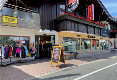 Ein Sportgeschäft mit einer großen Auswahl an Sportkleidung und Ausrüstung. Die Fassade ist mit Blumen dekoriert und es gibt ein Schild, das auf besondere Angebote hinweist.