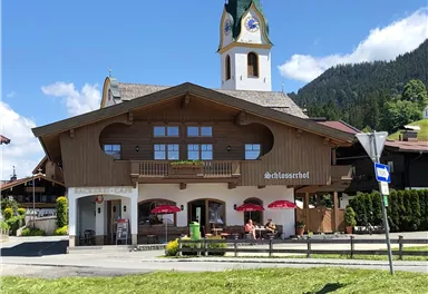A traditional alpine inn with a characteristic clock tower. In the foreground, there are tables with red umbrellas and a green meadow.