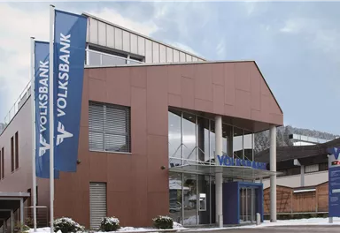 A modern bank building with large windows and two flags from the Volksbank. The surroundings are wintry with light snow.