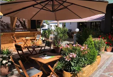 Ein einladender Gartenbereich mit Holzmöbeln und einem großen Sonnenschirm. Grünpflanzen und Blumen umgeben die Sitzplätze und schaffen eine angenehme Atmosphäre.