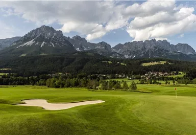 Eine malerische Golfplatzszene mit grünem Rasen und einem Sandbunker. Im Hintergrund erheben sich beeindruckende Berge unter einem bewölkten Himmel.
