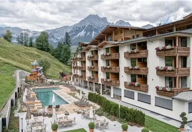 A modern hotel with wooden cladding and balcony flowers. In the background, majestic mountains and a swimming pool with loungers can be seen.