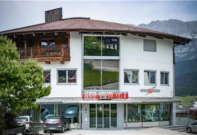 A modern building with an attractive, covered area. In the foreground, some cars and a green environment can be seen.