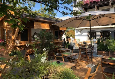 Ein einladender Außenbereich eines Restaurants mit Holzmöbeln und vielen Pflanzen. Sonnenschirme spenden Schatten an einem sonnigen Tag.