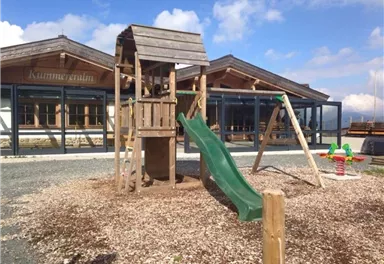 A playground with a wooden play tower, a green slide, and swings. In the background, modern buildings with large windows can be seen.