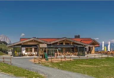 A cozy mountain restaurant with a large terrace. In the background, the Alps and a clear blue sky are visible.