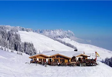 A cozy mountain cabin in the snow-covered Alps. Surrounded by snow-covered trees and a bright blue sky.