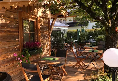 Eine gemütliche Terrasse vor einem Holzhaus mit Tischen und Stühlen. Umgeben von Pflanzen und Bäumen bietet sie eine einladende Atmosphäre.