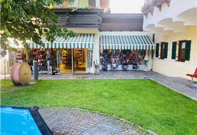 Ein Geschäft mit einem gestreiften Markisen, das bunte Waren anbietet. Im Vordergrund liegt eine grüne Rasenfläche und ein Pool.