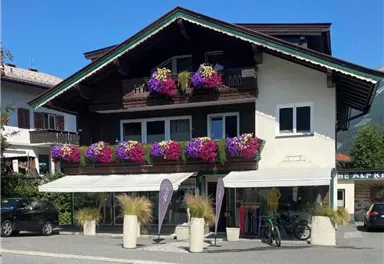 Ein charmantes Gebäude mit bunten Blumenbalkonen. Vor dem Haus stehen gleichmäßige Pflanzgefäße und einige Parkplätze.