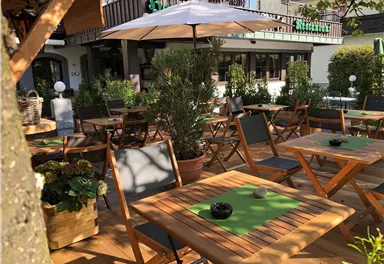 Ein gemütlicher Außenbereich mit Holztischen und Stühlen. Eine Sonne schützende Markise sorgt für Schatten in einer einladenden Atmosphäre.