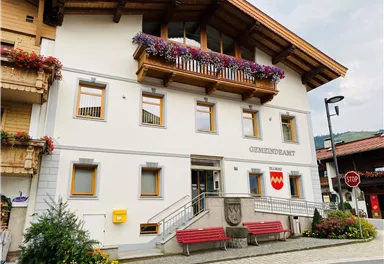 Ein schönes Gebäude mit einem Balkon, der mit Blumen geschmückt ist. Es handelt sich um das Gemeindeamt mit einer einladenden Treppe und zwei roten Bänken davor.