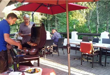 A sociable barbecue party outdoors. Two men are grilling meat on a terrace, surrounded by a beautiful garden.