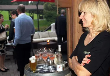 A server is standing at a table with drinks. In the background, several guests can be seen at an event.