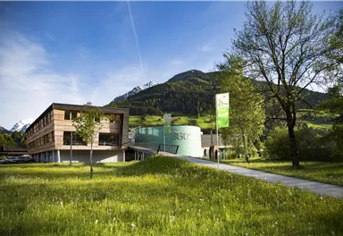 A modern building surrounded by green meadows and trees. In the background, mountains can be seen under a clear sky.