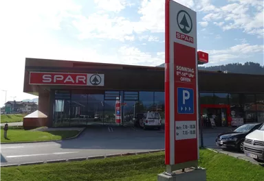 Ein SPAR Supermarkt mit großem Parkplatz und klarer blauer Himmel. Die Geschäftszeiten sind auf dem Schild angegeben.
