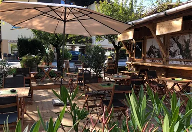 Eine gemütliche Terrasse mit Holzstühlen und Tischen unter einem großen Sonnenschirm. Rasen und Pflanzen umgeben den Sitzbereich und schaffen eine einladende Atmosphäre.