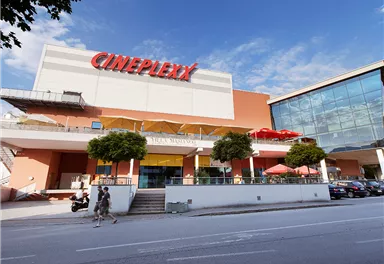 Ein modernes Kino mit der Aufschrift "Cineplexx" auf der Fassade. Vor dem Gebäude sind Menschen und Pflanzen zu sehen.