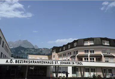 A modern hospital building named "A.O. District Hospital St. Johann in Tirol". Impressive mountains are visible in the background.