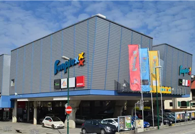A modern commercial building with the inscription "Jumex". There are several colorful flags and parking spaces in the foreground.
