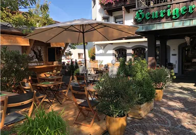 Ein einladendes Café mit Außenbereich, Tischen und Stühlen unter einem Sonnenschirm. Die Umgebung ist grün und freundlich gestaltet.