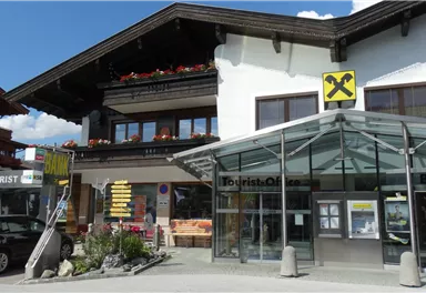 A modern building with a glass roof and an entrance to a bank. In the background, you can see a traditional chalet with flower boxes.