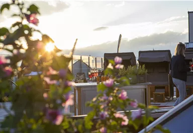 Eine Dachterrasse mit blühenden Pflanzen und einer Person, die einen Drink hält. Im Hintergrund ist ein schöner Sonnenuntergang zu sehen.