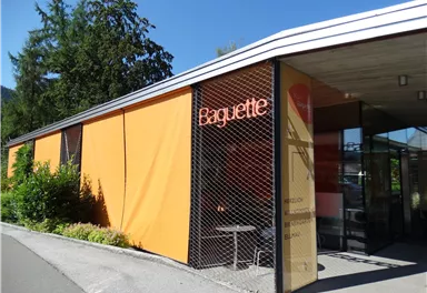 Ein modernes Gebäude mit einer auffälligen orangefarbenen Fassade und dem Schriftzug "Baguette". Umgeben von grünen Pflanzen und unter einem klaren blauen Himmel.