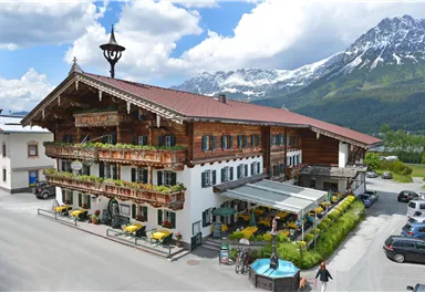 A charming alpine hotel with traditional wooden elements. In the background, impressive mountains and a blue sky can be seen.