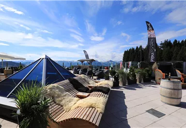 A cozy terrace with lounge furniture and plants offers a view of the mountains. The sky is clear and the atmosphere is inviting.