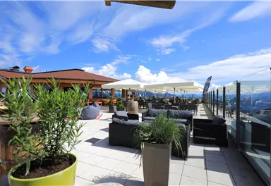 A cozy rooftop terrace with comfortable seating and sun umbrellas. The sky is blue with some clouds, and the view is impressive.