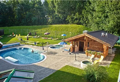 A beautiful outdoor area with a pool and hot tubs. In the background, there is a green meadow and a wooden hut.