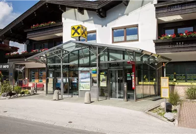 Eine moderne Bank mit einem großen Glasvordach. Sie befindet sich an einer belebten Straße und ist von gepflegten Pflanzen umgeben.