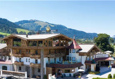 A charming hotel in alpine style with wooden cladding. The surroundings offer an impressive mountain backdrop and a green garden.