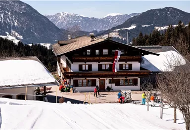 Ein traditionelles Alpenhaus liegt in einer schneebedeckten Landschaft. Skifahrer und Gäste sind auf dem Gelände unterwegs, während die Berge im Hintergrund sichtbar sind.