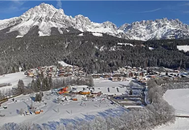 A picturesque winter landscape with snow-covered mountains and a small village. In the foreground, recreational activities in the snow can be seen.