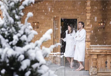 A couple in bathrobes stands in front of a wooden cabin in the snow. Thick snowflakes gently fall around them.