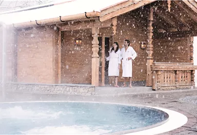 A cozy log cabin in the snow with a relaxing hot tub. Two people in bathrobes stand outside and enjoy the winter atmosphere.