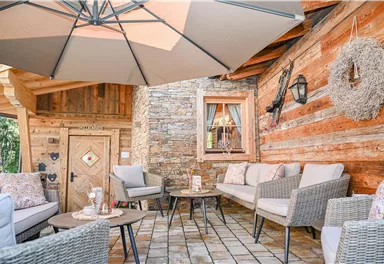 Eine gemütliche Sitzecke mit bequemen Möbeln unter einem großen Sonnenschirm. Die Wand besteht aus schönem Holz und Stein, was eine einladende Atmosphäre schafft.