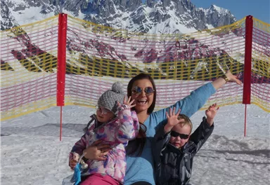 Eine fröhliche Frau sitzt mit zwei Kindern im Schnee und posiert. Im Hintergrund sind schneebedeckte Berge zu sehen.