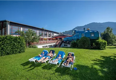 Eine entspannte Sommerlandschaft mit Sonnenliegen auf grünem Rasen. Im Hintergrund sind ein modernes Gebäude und ein blauer Traktor zu sehen.