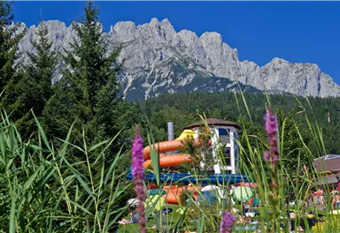 A water park with colorful slides and green meadows. In the background, majestic mountains and a clear blue sky can be seen.